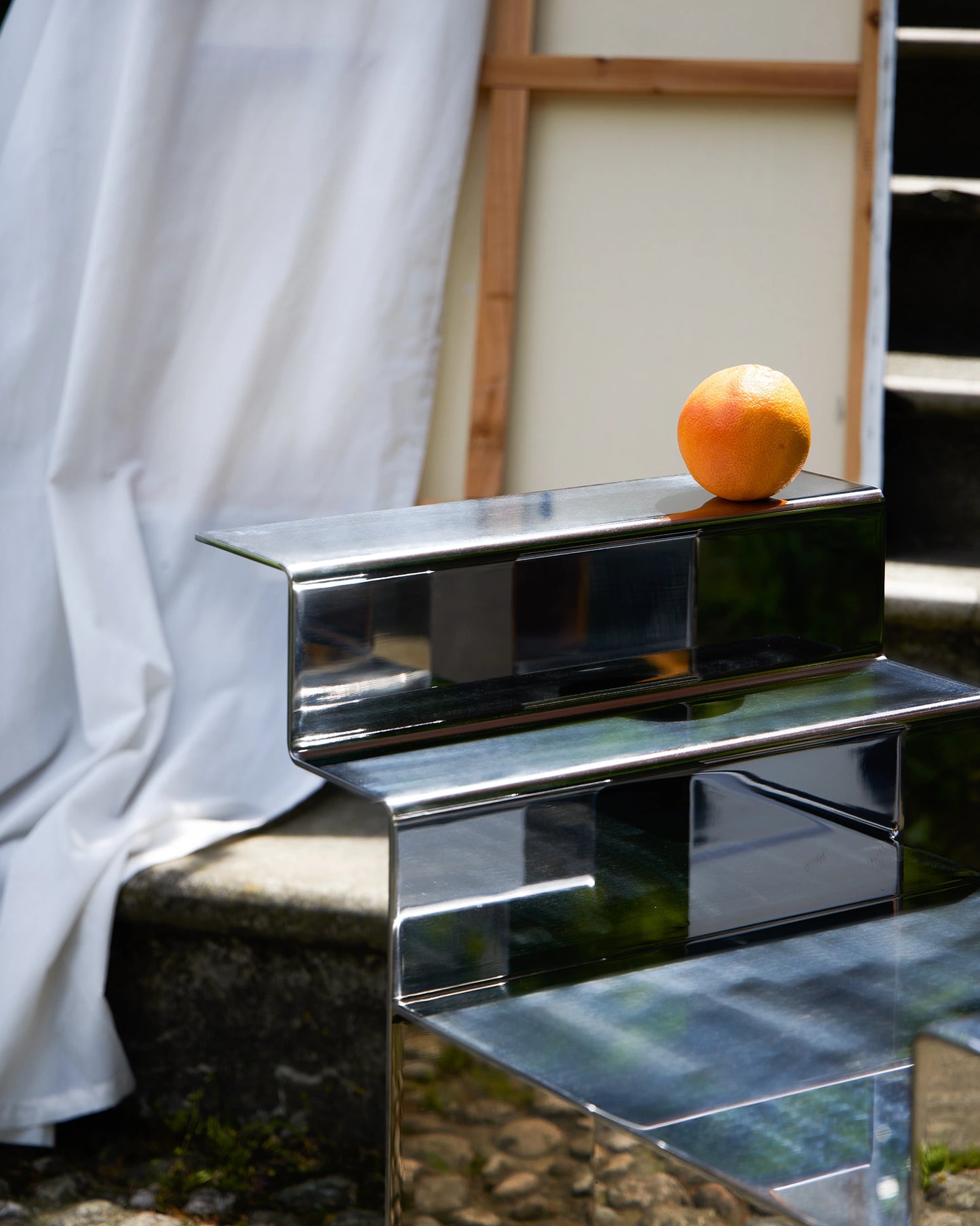 detail of a stainless-steel mirror chair with mesoamerican temple-like shape, with scene props like an orange and a curtain