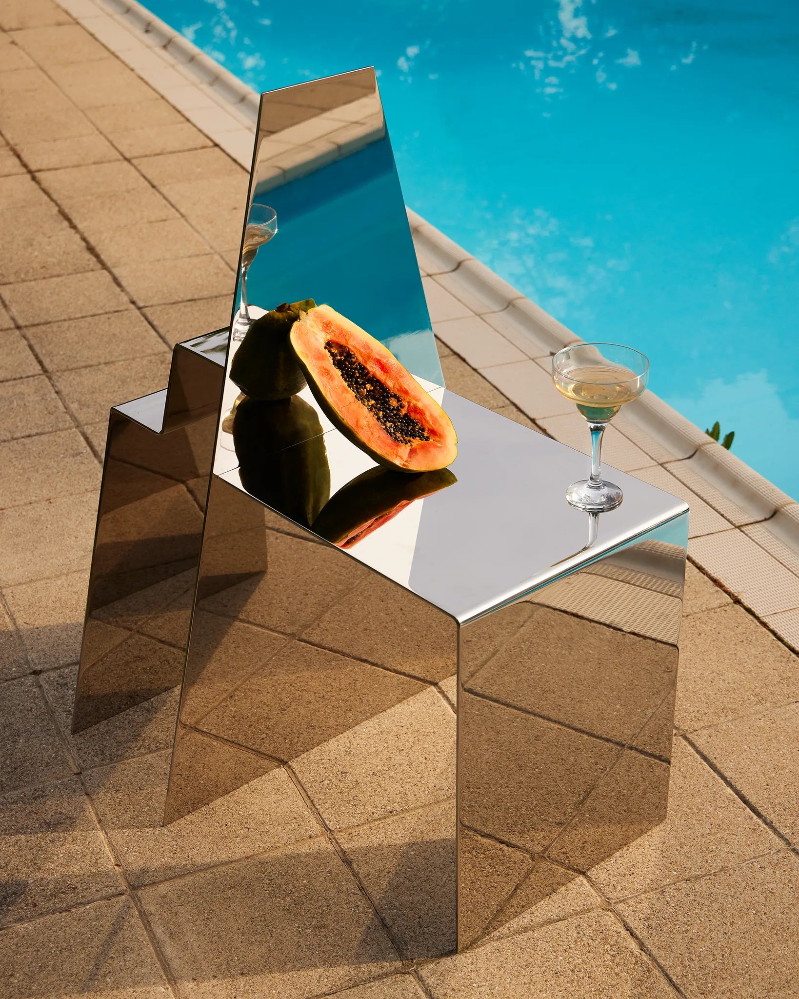 stainless-steel mirror chair with mesoamerican temple-like shape, in a summer setting on a poolside, with props like a half papaya and a glass of champagne