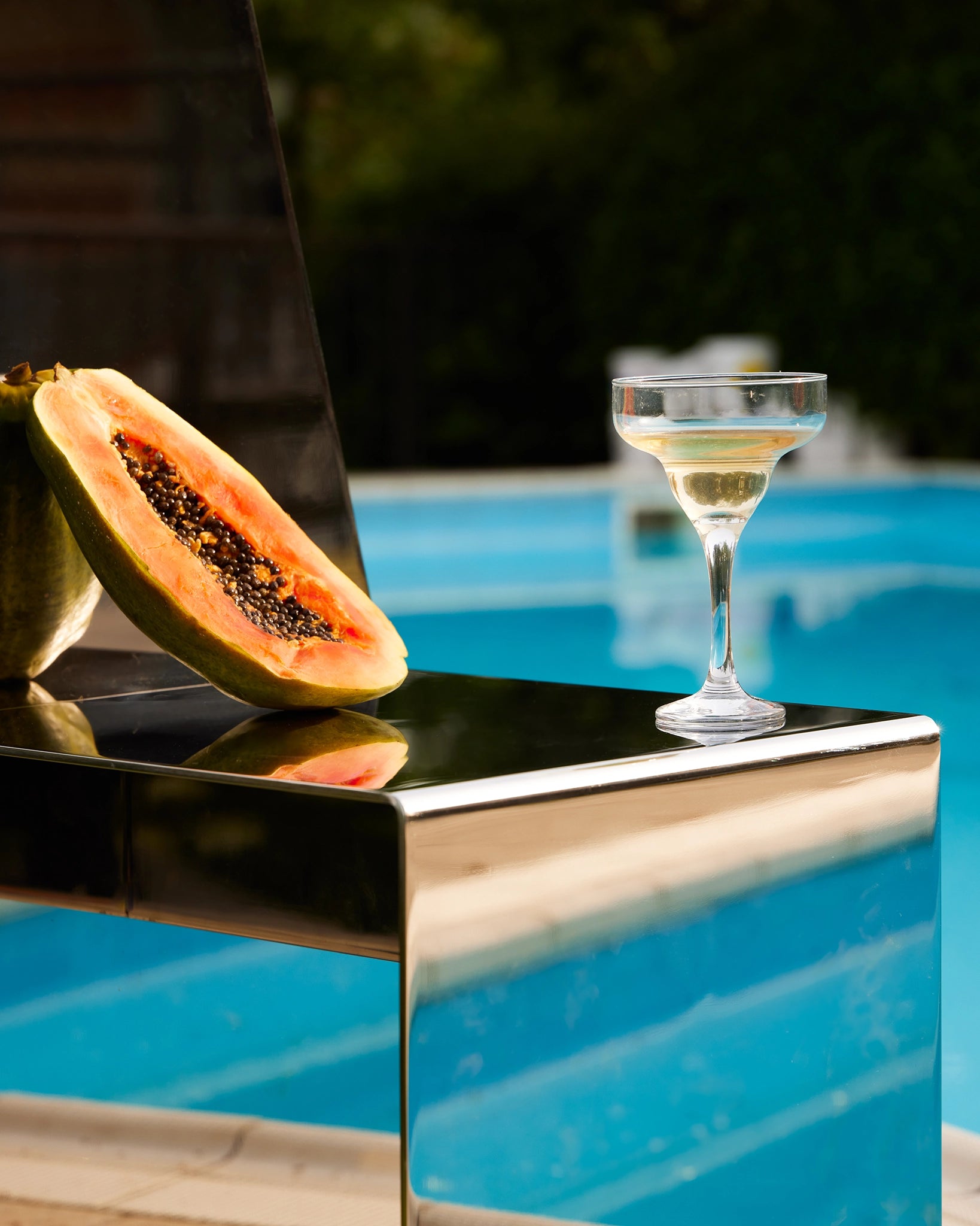 detail of a stainless-steel mirror chair with mesoamerican temple-like shape, in a summer setting on a poolside, with props like a half papaya and a glass of champagne