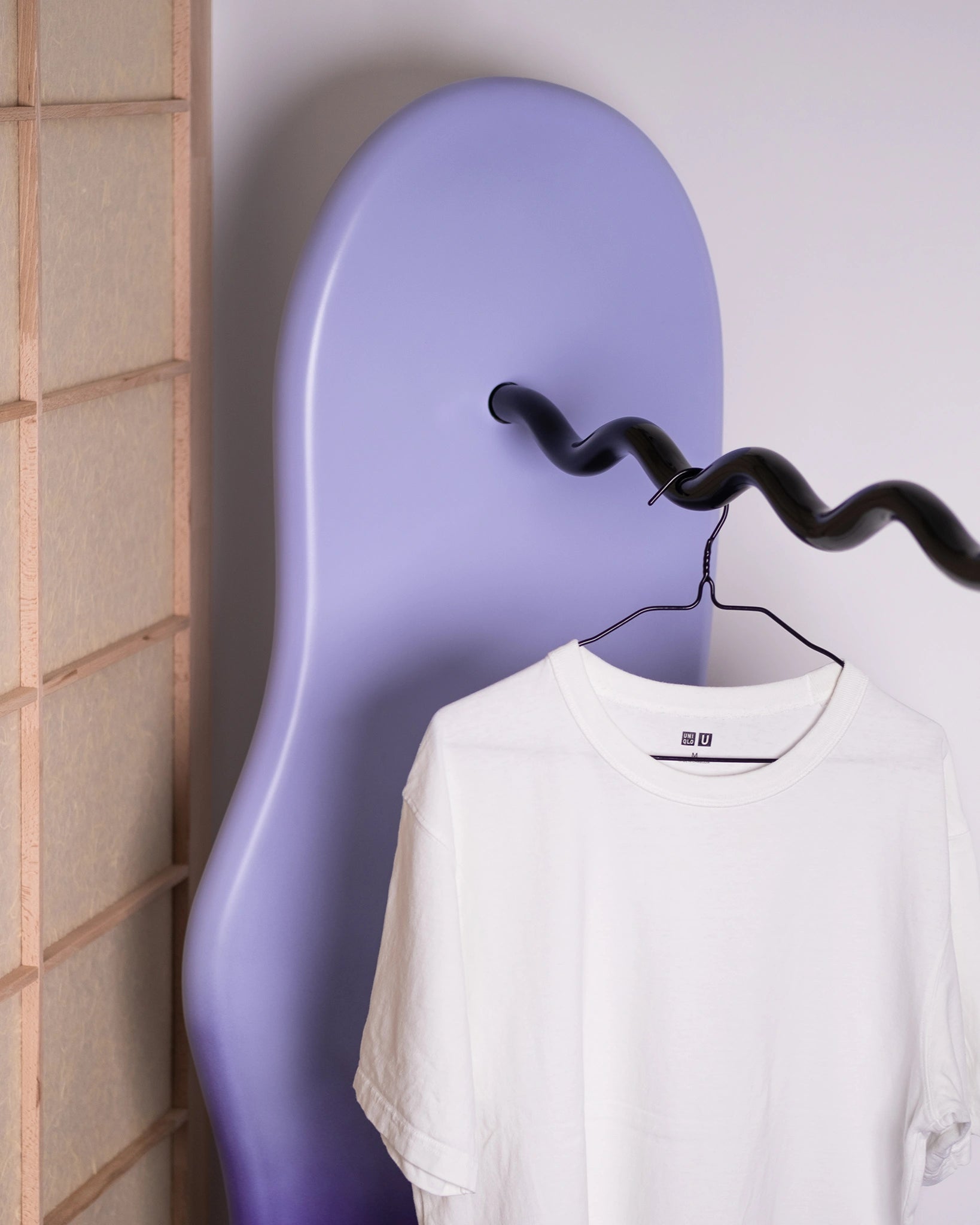 detail of a clothing rack in a home setting with soft shapes and a wavy metal hanging tube, purple gradient, t-shirt on it